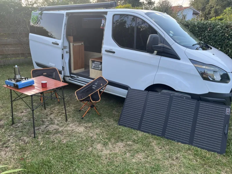 Peggy, a white compact van, with a folding table, stove, chairs and a portable solar panel set up beside it