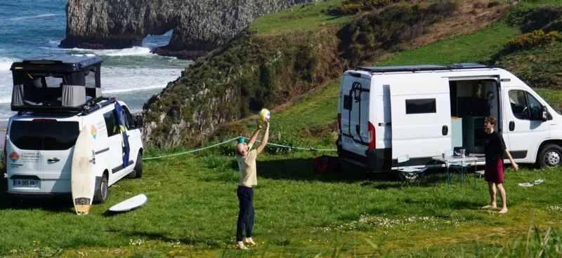 Les deux vans RB-CapSO, Pénélope et Peggy, garés sur une falaise verte au-dessus de l'océan, des voyageurs et une planche de surf à côté