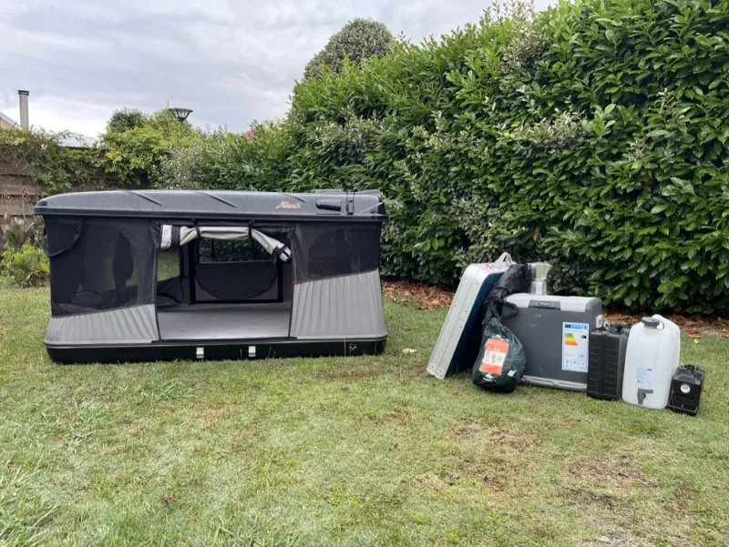 The hard-shell roof tent, open on a lawn, next to a cool box, a battery and a water container