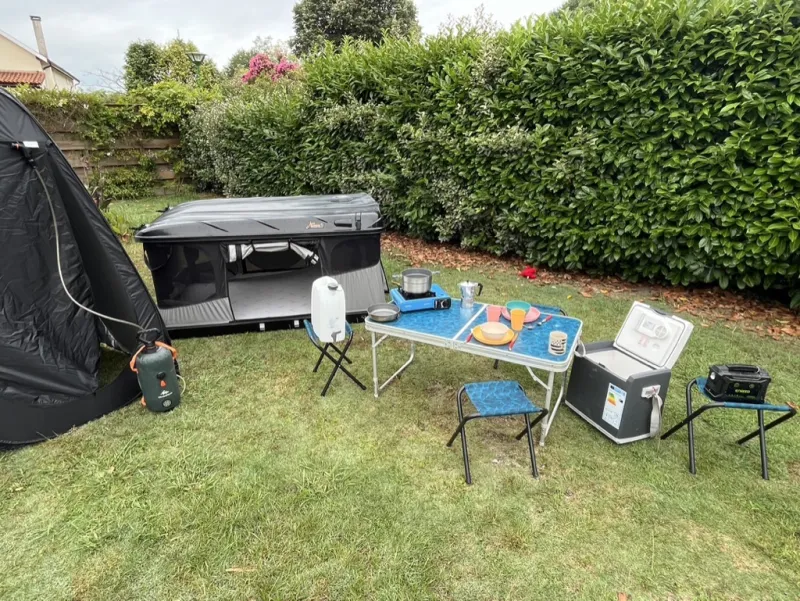 Le matériel de camping déplié devant la tente de toit : table, tabourets, réchaud, glacière, bidon d'eau, batterie et cabine de douche